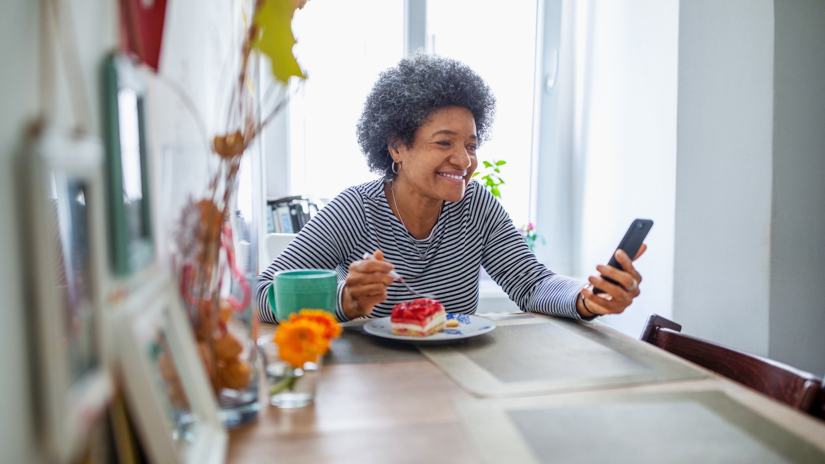 Ältere Dame sitzt am Esstisch, geniesst ihr Essen und hält in der anderen Hand ein Smartphone und lächelt darauf